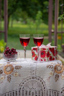 Kırmızı bir içecek ile iki bardak, kristal vazo içinde olgun bir kiraz, bahçede güzel bir masa örtüsü ile dekore edilmiş bir masa üzerinde bir hediye standı ile parlak bir kutu.