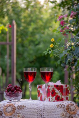 Kırmızı bir içecek ile iki bardak, kristal vazo içinde olgun kiraz, çiçek açan gül ler yakın sokakta güzel bir masa örtüsü ile dekore edilmiş bir masada bir hediye standı ile parlak bir kutu.