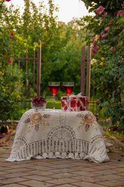 Kırmızı bir içecek ile iki bardak, kristal vazo olgun kiraz, tırmanma güller arasında bahçede güzel bir masa örtüsü ile dekore edilmiş bir masa üzerinde bir hediye standı ile parlak bir kutu.