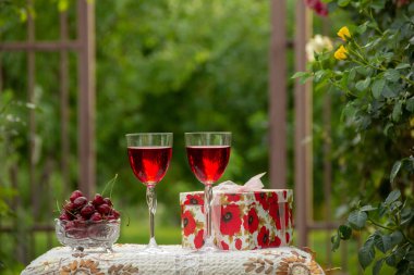 Kırmızı bir içecek ile iki bardak, kristal vazo içinde olgun kiraz, çiçek açan gül ler yakın sokakta güzel bir masa örtüsü ile dekore edilmiş bir masada bir hediye standı ile parlak bir kutu.
