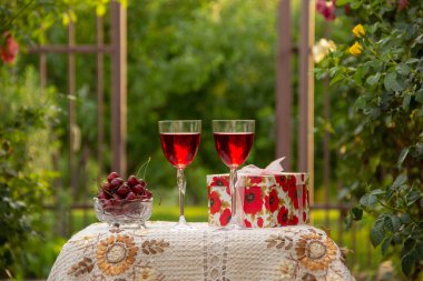 Kırmızı bir içecek ile iki bardak, kristal vazo içinde olgun kiraz, çiçek açan gül ler yakın sokakta güzel bir masa örtüsü ile dekore edilmiş bir masada bir hediye standı ile parlak bir kutu.