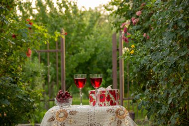 Kırmızı bir içecek ile iki bardak, kristal vazo olgun kiraz, tırmanma güller arasında bahçede güzel bir masa örtüsü ile dekore edilmiş bir masa üzerinde bir hediye standı ile parlak bir kutu.