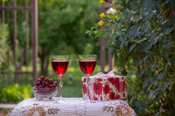 Kırmızı bir içecek ile iki bardak, kristal vazo içinde olgun kiraz, çiçek açan gül ler yakın sokakta güzel bir masa örtüsü ile dekore edilmiş bir masada bir hediye standı ile parlak bir kutu.