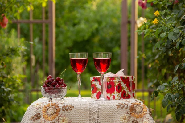 Kırmızı bir içecek ile iki bardak, kristal vazo içinde olgun kiraz, çiçek açan gül ler yakın sokakta güzel bir masa örtüsü ile dekore edilmiş bir masada bir hediye standı ile parlak bir kutu.