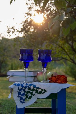 İki cam mavi bardak, iki kitap, bir elma ve bir tabure üzerinde çiçek bir masa örtüsü ile süslenmiş, bir ağaç üzerinde olgun bir armut ile yaz bahçesi, gün batımı.