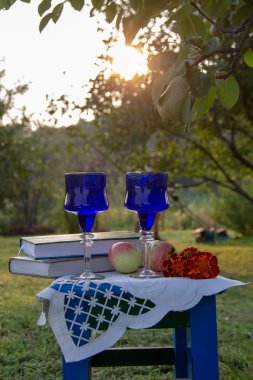İki cam mavi bardak, iki kitap, bir elma ve bir tabure üzerinde çiçek bir masa örtüsü ile süslenmiş, bir ağaç üzerinde olgun bir armut ile yaz bahçesi, gün batımı.