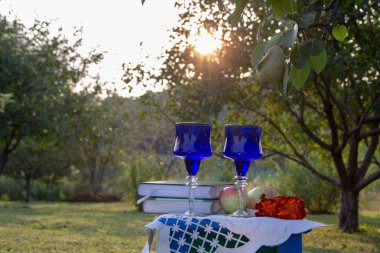 İki cam mavi bardak, iki kitap, bir elma ve bir tabure üzerinde çiçek bir masa örtüsü ile süslenmiş, bir ağaç üzerinde olgun bir armut ile yaz bahçesi, gün batımı.
