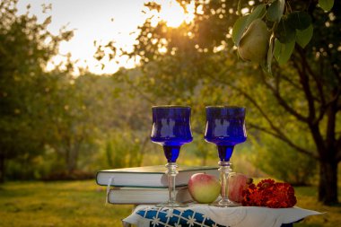 İki cam mavi bardak, iki kitap, bir elma ve bir tabure üzerinde çiçek bir masa örtüsü ile süslenmiş, bir ağaç üzerinde olgun bir armut ile yaz bahçesi, gün batımı.