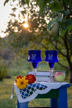 İki cam mavi bardak, iki kitap, bir elma ve bir tabure üzerinde çiçek bir masa örtüsü ile süslenmiş, bir ağaç üzerinde olgun bir armut ile yaz bahçesi, gün batımı.