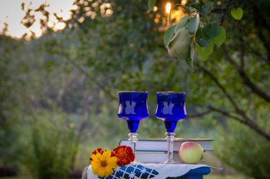 İki cam mavi bardak, iki kitap, bir elma ve bir tabure üzerinde çiçek bir masa örtüsü ile süslenmiş, bir ağaç üzerinde olgun bir armut ile yaz bahçesi, gün batımı.