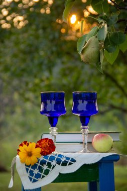 İki cam mavi bardak, iki kitap, bir elma ve bir tabure üzerinde çiçek bir masa örtüsü ile süslenmiş, bir ağaç üzerinde olgun bir armut ile yaz bahçesi, gün batımı.