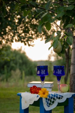 İki cam mavi gözlük, iki kitap, bir elma ve çiçekler bir tabure üzerinde bir masa örtüsü ile süslenmiş standı, bir yaz bahçesi ve olgun bir armut ile bir ağaç etrafında.