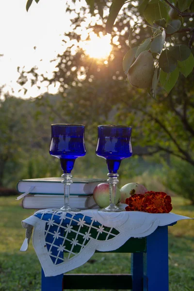 İki cam mavi bardak, iki kitap, bir elma ve bir tabure üzerinde çiçek bir masa örtüsü ile süslenmiş, bir ağaç üzerinde olgun bir armut ile yaz bahçesi, gün batımı.