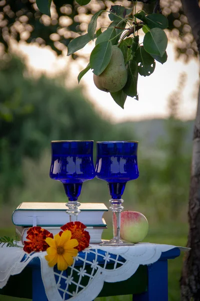 İki cam mavi gözlük, iki kitap, bir elma ve çiçekler bir tabure üzerinde bir masa örtüsü ile süslenmiş standı, bir yaz bahçesi ve olgun bir armut ile bir ağaç etrafında.