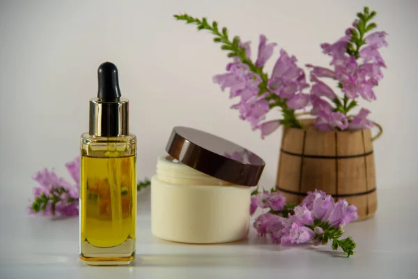 Glass bottle with golden natural face oil and a dropper, packaging for face cream, a fragrant bouquet of purple flowers in a wooden barrel on a white background.