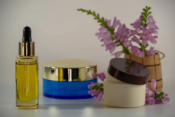 Glass jar with a pipette and natural face oil, two packs with face cream, a wooden barrel with a fragrant bouquet of flowers on a white background.