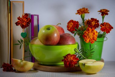 Yeşil bir tabakta olgun elma natürmort, küçük bir dekoratif kova içinde kadife bir buket, kitaplar, beyaz bir arka plan üzerinde bir gülümseme şeklinde bir şiş ile elma yarıya.