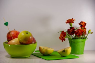 Yeşil bir tabakta olgun elma, küçük bir yeşil dekoratif kova, kesme tahtası, bir gülümseme ile bir şiş, beyaz bir arka plan üzerinde ikiye kesilmiş bir elma çiçekaçan turuncu kadife çiçeği bir buket.