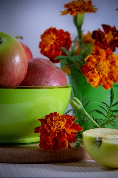 Ripe apples in a green plate, a bouquet of blooming orange marigolds in a small green decorative bucket, half apple shaped smiley on a white background.