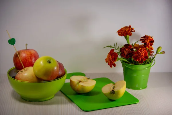 Ripe apples in a green plate, a bouquet of blooming orange marigolds in a small green decorative bucket, a cutting board, a skewer with a smile, an apple cut in half on a white background.