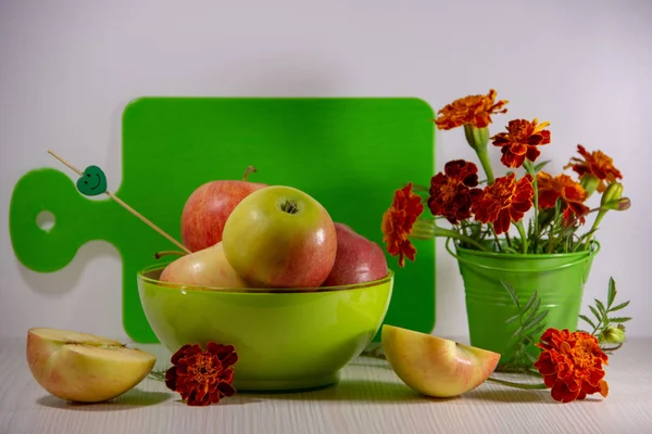 Ripe apples in a green plate, a bouquet of blooming orange marigolds in a small green decorative bucket, a cutting board, a skewer with a smile, an apple cut in half on a white background.
