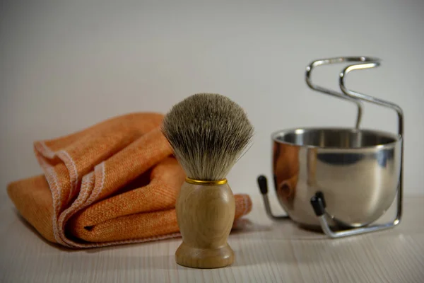 Shaving accessories: a wooden shaving razor with a natural bristle brush, a metal bowl on a stand, an orange towel. Light background.