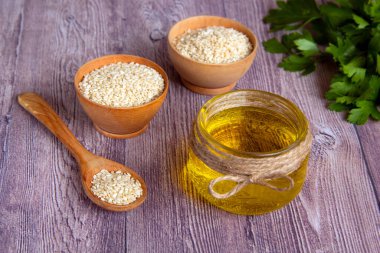 Cam kavanoz susam yağı ahşap bir kâse ve kaşıkta çiğ beyaz susam tohumu ahşap arka planda taze yeşillikler. Sağlıklı bir yaşam tarzı. Doğal ve sağlıklı yağ.