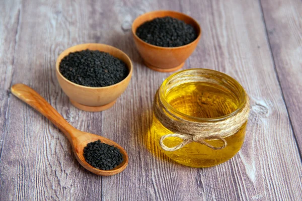 Glass jar of sesame oil and raw black sesame seeds in a wooden bowl and spoon on a wooden background.Useful natural oil. Natural and healthy food concept.