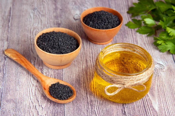 Glass jar of sesame oil, raw black sesame seeds in a wooden bowl and spoon, fresh greens on a wooden background. Healthy lifestyle. Natural and healthy oil.