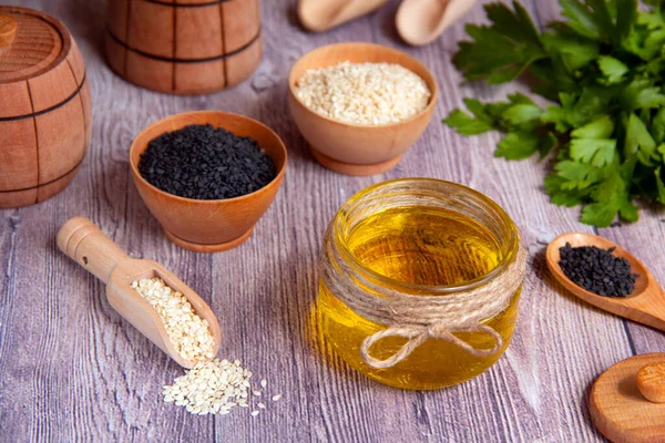 Glass jar of sesame oil, raw white and black sesame seeds, wooden dish, fresh herbs on a wooden background. Healthy lifestyle. Natural and healthy oil.