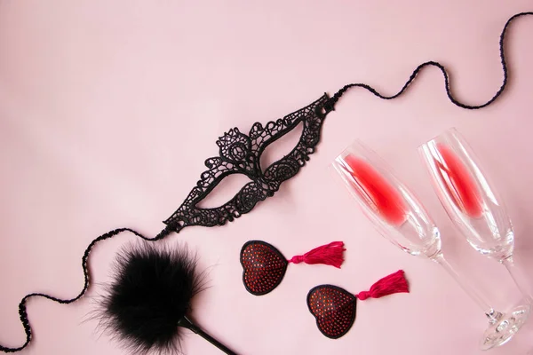 Bir kadın için dantel maske, kırmızı püsküllü burlesk köfteler, bir tüy küreği, pembe arka planda bir kadeh şarap. Yetişkinler için seksi oyuncaklar. Düz yatış, üst görünüm.