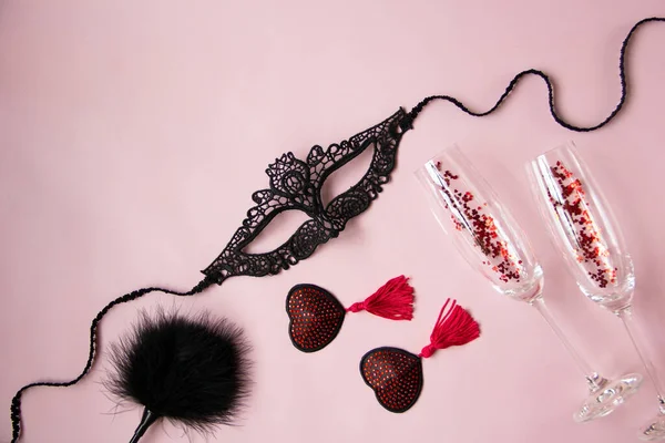Bir kadın için dantel maske, kırmızı püsküllü burlesk köfteler, bir tüy küreği, pembe arka planda bir kadeh şarap. Yetişkinler için seksi oyuncaklar. Düz yatış, üst görünüm.