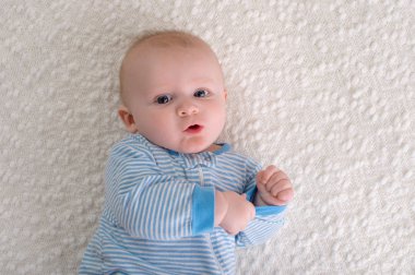 Beyaz bir battaniye sırtında yatarken 2 aylık erkek bir bebek. O mavi ve beyaz çizgili pijama giyiyor ve kameraya bakıyor.