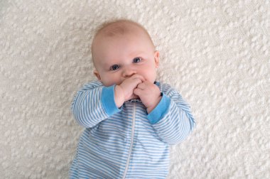 Beyaz bir battaniye sırtında yatarken 2 aylık erkek bir bebek. O mavi ve beyaz çizgili pijama giyiyor ve elini ağzına ve kameraya bakıyor.