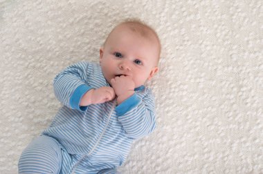 Beyaz bir battaniye sırtında yatarken 2 aylık erkek bir bebek. O mavi ve beyaz çizgili pijama giyiyor ve elini ağzına ve kameraya bakıyor.