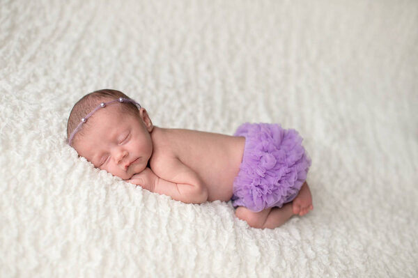 Portrait of a sleeping, 2 week old newborn baby girl wearing frilly, lavender purple bloomers and a pearl headban. Shot in the studio on a white blanket.