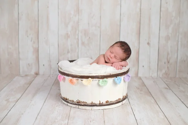 一个两周大的新生女婴睡在一个小木桶里 有一颗彩虹色的心花环 拍摄在工作室的白色 木质背景 — 图库照片