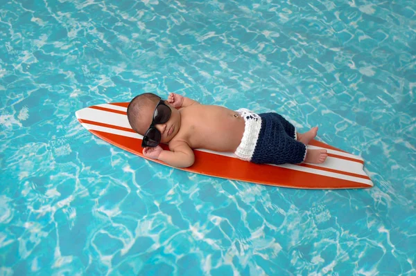 九天的初生婴儿男孩睡在一个小 橙色和白色的冲浪板 他穿着深蓝 钩编的短裤和太阳镜 — 图库照片