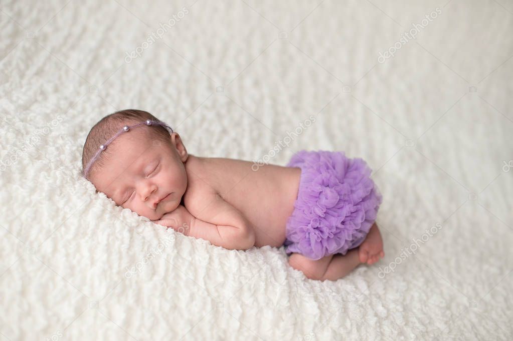 Portrait of a sleeping, 2 week old newborn baby girl wearing frilly, lavender purple bloomers and a pearl headban. Shot in the studio on a white blanket.