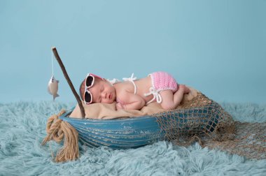 Dokuz gün eski bir yeni doğmuş bebek kız portresi. O bir minyatür balıkçı teknesi uyku ve güneş gözlüğü ve pembe bikini giyiyor. Studio bir aqua renkli flokati halı üzerinde ateş.