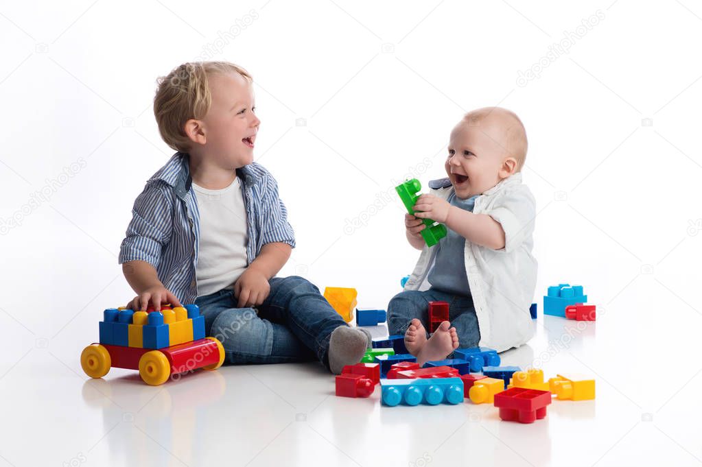 A two year old boy playing with his seven month old baby brother playing with building blocks. Shot in the studio on a white, seamless backdrop.