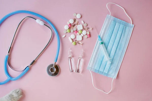 Veterinary medicine concept. Cat's paw, pills, medical mask and phonendoscope on a pink background