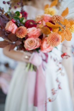 Gelin, elinde turuncu, kırmızı çiçekler ve böğürtlenlerle güzel bir sonbahar buketi tutuyor. Gelinin elinde kurdelelerle sonbahar buketi