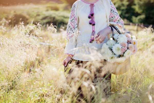 Woman in ukrainian traditional shirt holds bouquet of white and pink chrysanthemums in straw bag. Autumn concept, walk in the field at sunlight with flowers