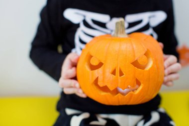 İskelet kostümlü beyaz çocuk kambur bir balkabağı tutuyor ve Cadılar Bayramı yemeği ve süslemeleriyle masada oturuyor.