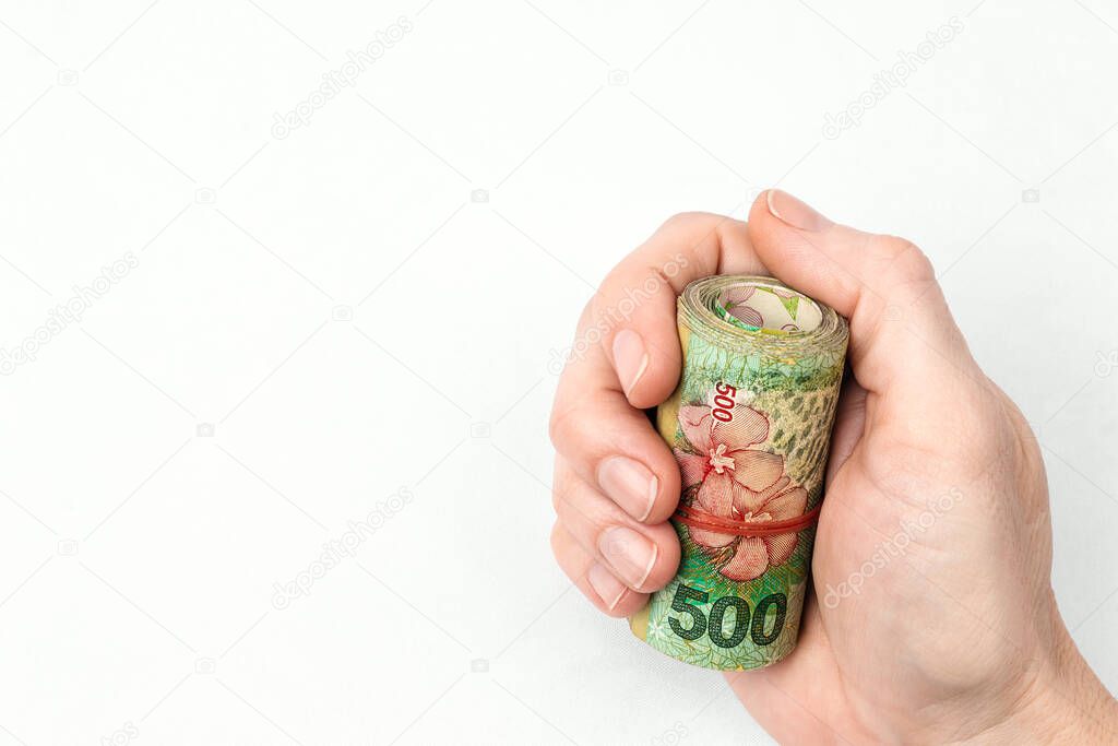 Female hand of white Caucasian skin and short nails without enamel holds in her closed fist a bundle of 500 Argentine pesos bills rolled up and tied with red elastic band on white background.