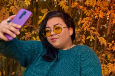 Bir kız sonbahar ağaçlarının arka planında selfie çekiyor. Cep telefonundaki fotoğraf.