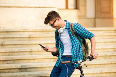 gözlük ve onun bisiklet Üniversitesi önünde sokakta duran ve cep telefonu, seyir Bisiklet ile gülümseyerek, sosyal medya kontrol genç öğrenci