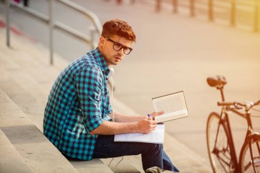 okul veya üniversite merdivenlerde durup, bir kitap okuma ve not alma ödevini bakarak gözlüklü genç öğrenci