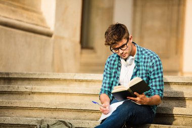 okul veya üniversite merdivenlerde durup, bir kitap okuma ve not alma ödevini bakarak gözlüklü genç öğrenci
